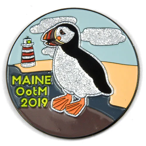 Custom trading pin featuring a puffin with glittery white feathers standing on a coastal cliff, with a red and white striped lighthouse in the background. The pin includes the text “MAINE OoTM 2019,” representing a 2019 Odyssey of the Mind team from Maine.