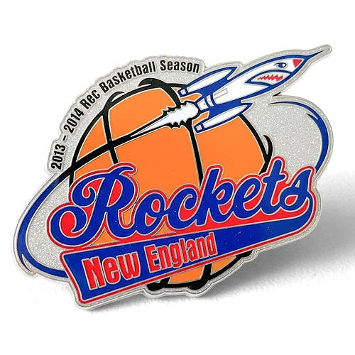 New England Rockets custom trading pin from the 2013–2014 rec basketball season. Custom trading pin for the New England Rockets basketball team, featuring a rocket shooting through an orange basketball with a silver glitter background. The pin includes the text “2013–2014 Rec Basketball Season” and bold red and blue lettering.