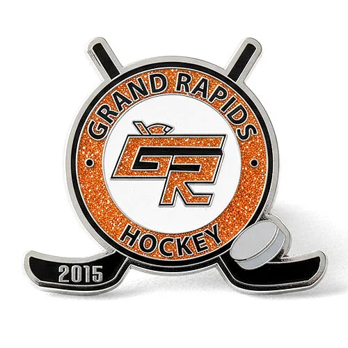 Grand Rapids Hockey 2015 custom trading pin with glitter and GR logo. Custom trading pin for Grand Rapids Hockey, featuring a circular design with glittery orange accents, a stylized “GR” logo in the center, and two black hockey sticks crossed behind the pin. A gray puck and the year “2015” appear at the base.